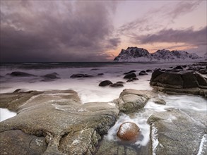 Rounded rocks on Utakleiv beach in a dramatic cloudy atmosphere, snow-capped mountains in the