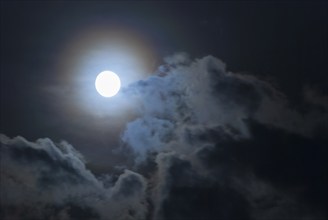 The full moon shines brightly in a cloudy, stormy, black sky and has a halo in rainbow colours