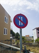 Red traffic sign with blue and white no parking symbol in front of buildings and a clear sky, no