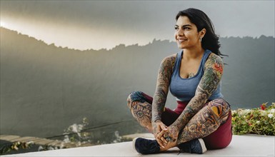 Satisfied woman with colourful tattoos sits smiling in front of a mountain landscape, AI generated,