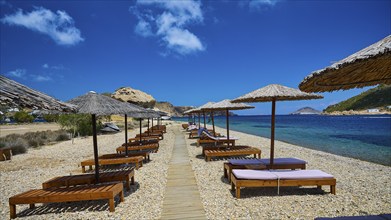 A beach with a wooden walkway and deckchairs under thatched umbrellas, the view of the clear water