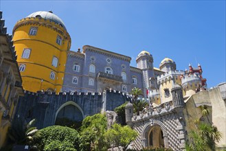 Castle with grey and yellow towers against a blue sky, surrounded by plants, Palácio Nacional da