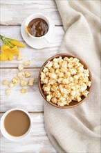 Popcorn with caramel in wooden bowl and a cup of coffee on a white wooden background and linen