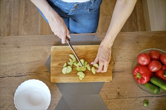 Overhead view of female hands slicing fresh cucumber on wooden chopping board