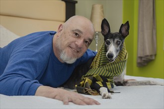 Mature man posing and smiling while looking at the camera with his greyhound pet on the bed