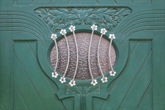 Close-up of a green door with elaborate decorations and a small oval glass window with a metal