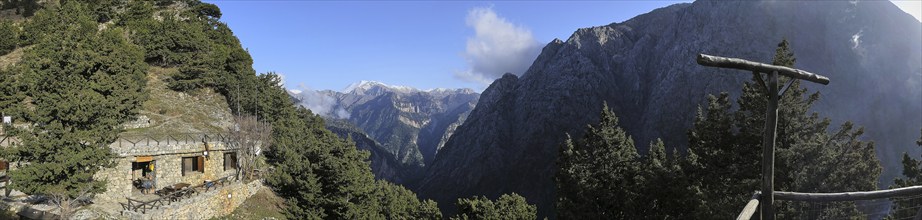 Mountain hut nestled in a wooded mountain landscape with views of snow-capped peaks under a blue