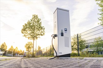 An electric charging station on a cobblestone street at sunset, surrounded by trees and urban