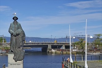 Statue of a fisherman in front of a river and a bridge under a blue sky, the last Viking,