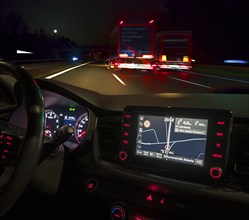 Night-time road scene with two lorries, in front an illuminated cockpit with sat nav of a car,