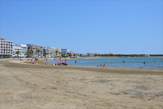 A sunny, sandy seafront promenade with people, palm trees and modern buildings along the coast, Le