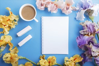 Purple, pink, yellow iris flowers and a cup of coffee with notebook on pastel blue background.