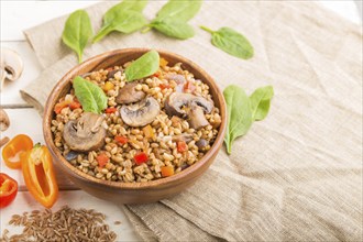 Spelt (dinkel wheat) porridge with vegetables and mushrooms in wooden bowl on a white wooden