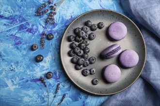 Purple macarons or macaroons cakes with blueberries on ceramic plate on a blue concrete background