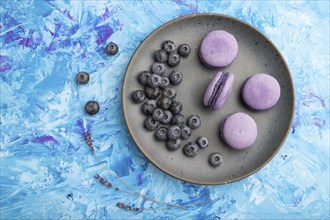 Purple macarons or macaroons cakes with blueberries on ceramic plate on a blue concrete background.