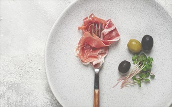 Prosciutto on a fork, top view, olives and micro-greens on a plate, light background, no people