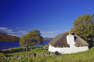 Small white house with thatched roof by the lake, surrounded by meadows and trees under a blue sky,