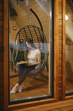 Young woman sitting in a cozy hanging chair, engrossed in reading a book. A woman spends her