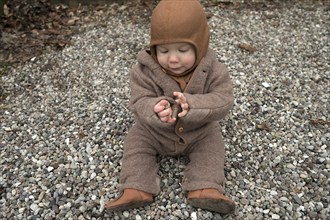 Small child, 8 months, wrapped up warm, sitting on the gravel and playing with stones,