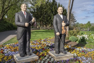 Two statues of men in classic suits stand side by side in a flowering garden under a blue sky,