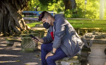 Man with a mask sitting on a bench with his phone, young man with a mask sitting with a cell phone,