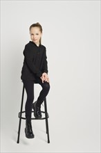 Young girl dressed in a black shirt and tights sits casually on a high stool against a white