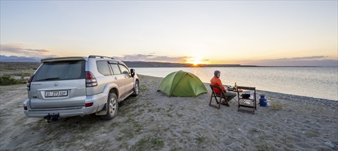 Camping at the lakeside, young man sitting next to tent on camping chair, having dinner, off-road