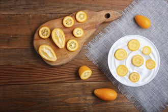 Greek yogurt with kumquat pieces in a white plate on a brown wooden background, top view, flat lay,