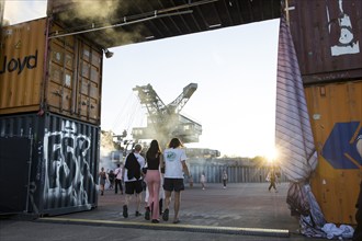 Festival visitors walk in the evening sun through a bridge made of containers in front of one of