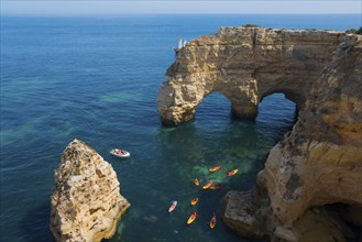 Stone arches and rocks jut out of the clear blue sea, kayaks and a small boat glide across the