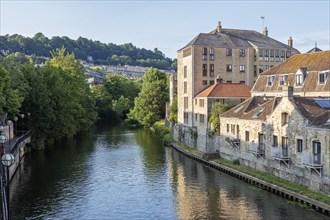 River flows quietly next to historic buildings and green trees in the sunlight, Bath