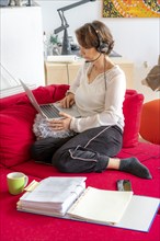 Woman, mid-50s, works from home, with laptop and communicates with colleagues via headset, home