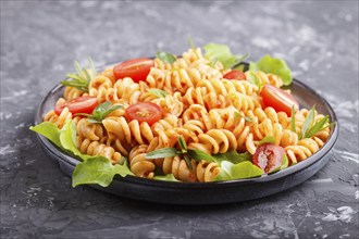 Fusilli pasta with tomato sauce, cherry tomatoes, lettuce and herbs on a black concrete background.