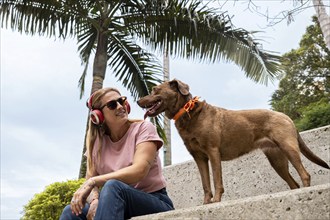 A woman, wearing red headphones and sunglasses, sits while looking at her pet with palm trees in