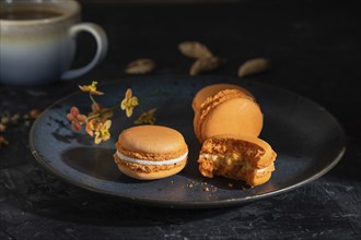 Orange macarons or macaroons cakes with cup of coffee on a black concrete background. Hard light,