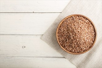 Wooden bowl with unpolished brown rice on a white wooden background and linen textile. Top view,