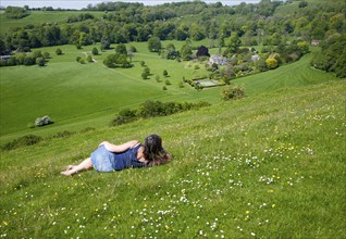 Woman lying on chalk grassland looking into deep green valley containing Rainscombe House, Oare