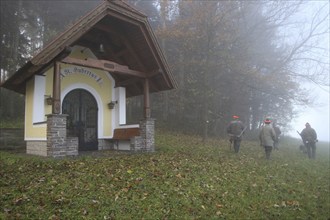 Hunters, in light autumn fog, pass Hubertus Chapel to hunt hares (Lepus europaeus) Lower Austria,