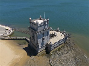 A historic tower by the sea, standing on a rocky sandy beach, with blue sky above, aerial view,