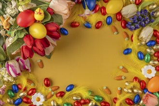 Colourful Easter frame with spring flowers, feathers, chocolate eggs and Easter eggs on a yellow