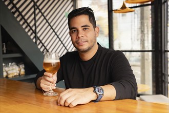 Stylish Latin man dressed in casual clothes enjoys a glass of beer sitting at the bar of a modern