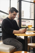 Young Latino man sitting at a bar table, using his cell phone while enjoying a glass of craft beer.