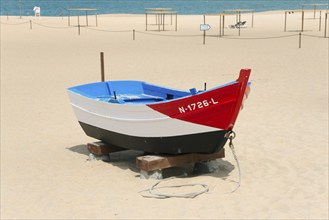 A small red, blue and white boat on a sandy beach, tied up with a rope, Praia da Nazare beach,