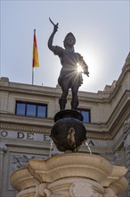 Sunlight shines behind a statue on a fountain, a flag flies in the background, Seville