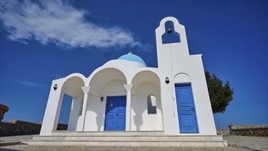 White church with blue doors and a bell in front of a blue sky and tree, Church of Profitis Ilias,