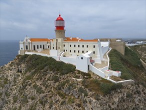 A red lighthouse and white buildings stand on a cliff, surrounded by vegetation and the ocean,