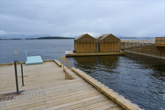 Outdoor sea pool by the fjord with small wooden sauna huts, overcast sky and calm water, Molde,