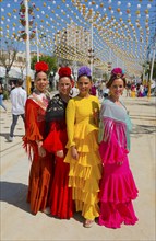 Four woman in bright flamenco dresses and flowers in their hair walk along a festival street under