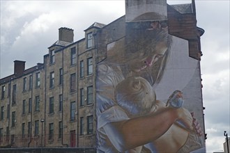 A mural shows a woman holding a baby and a bird in her arms. A building can be seen in the