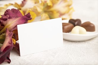 White business card with chocolate candies and iris flowers on gray concrete background. side view,